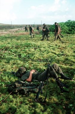 A Seabee from Naval Mobile Construction Battalion 3 (NMCB-3), his helmet over his face, rests after a day on the firing range during Exercise Kennel Bear '89. Marines from 3rd Force Service Support Group (3rd FSSG) walk by in the background