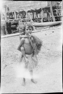 Child carrying an infant in front of a canoe amongst house poles, Papua, ca. 1923 / Sarah Chinnery