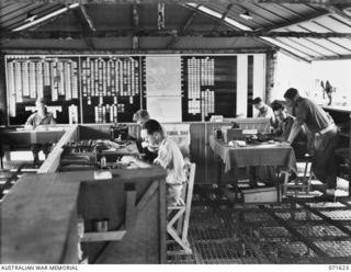 LAE, NEW GUINEA. 1944-03-24. THE ORDERLY ROOM AND ADMINISTRATION STAFF AT WORK IN THE AUSTRALIAN FORTRESS WORKSHOP. THE WALL TO THE REAR HOLDS INFORMATION ON ESTABLISHMENT, MANNING, AREA MAPS AND ..