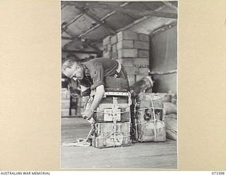 NADZAB, NEW GUINEA. 1944-05-25. VX48166 CORPORAL R N BEATTY, NON COMMISSIONED OFFICER IN CHARGE OF PACKING AT THE 3RD AIR MAINTENANCE COMPANY (1), ADJUSTS ROPES ON A SMALL ARMS AMMUNITION PACK. ON ..