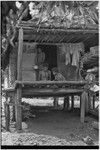 Family scene: man and child with a book, woman with tea kettle in doorway of small house