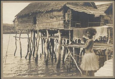 Woman wearing a grass skirt with nets drying and village houses behind, Hanuabada, Papua, 1921 / Sarah Chinnery