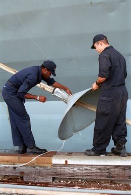 Two US Navy (USN) Sailors assigned to the USS BLUE RIDGE (LCC-19) attach rat guards to the mooring lines, while the ship is docked at Sierra Pier, Guam during Exercise TANDEM THRUST 99