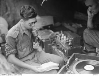 Pearl Ridge, Bougainville. 1945-03-07. QX61571 Private (Pte) J. Bavier speaking into a microphone, with VX19520 Signalman J. Ailes looking on. Both are from the Far Eastern Liaison Office (FELO) ..