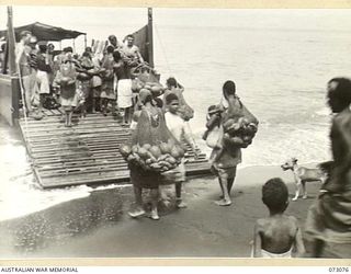YAMBO, NEW GUINEA, 1944-05-12. NATIVES LOAD A BARGE WITH PRODUCE FROM NATIVE GARDENS. THE "BILLUM" (STRING BAG), HUNG FROM THE HEAD, ASSISTS THE CARRYING OF PRODUCE. THE BARGE, CHARTERED FROM LAE ..