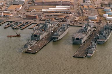 Aerial view, looking north-northeast, of a section of the Norfolk Naval Base showing piers 5 and 6. Tied up at the left is a large number of yard tug boats (YTBs). Tied on the north side of pier 6 is the amphibious assault ship USS Nassau (LHA-4) and the guided missile frigate USS Robert G. Bradley (FFG-49) with the decommissioned destroyer tender Puget Sound (AD-38) moored to the south side. The amphibious assault ship USS Saipan (LHA-2) is moored on the north side of pier 5 with the fast combat support ship USS Supply (AOE-6) and the Naval Reserve guided missile frigate USS Escotin (FFG-15) moored on the south side