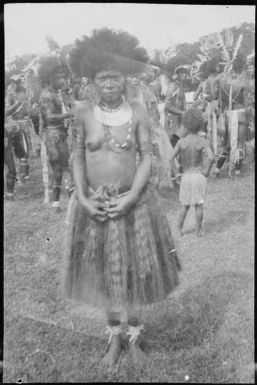 Woman wearing a grass skirt standing in front of dancers, Papua, ca. 1923 / Sarah Chinnery