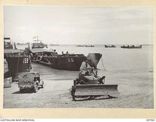 WEWAK BEACH, NEW GUINEA. 1945-10-18. A BULLDOZER PULLING LANDING CRAFT, TANK UP ONTO BEACH. AS THE BARGE IS UNLOADED THE LOSS OF WEIGHT TENDS TO MAKE IT FLOAT OFF THE BEACH. THE BULLDOZER KEEPS THE ..