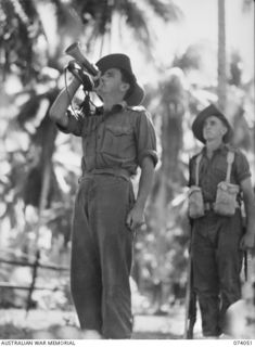 SIAR, NEW GUINEA. 1944-06-20. VX137515 CORPORAL G.T. MILLS (1) SOUNDS "REVEILLE" ON HIS BUGLE WHILE Q136596 PRIVATE H.W.L. DYER (2) STAND TO ATTENTION AT HEADQUARTERS, 58/59TH INFANTRY BATTALION