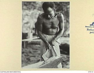 TSIMBA AREA, BOUGAINVILLE ISLAND. 1945-02-19. NEW GUINEA ADMINISTRATIVE UNIT (NGAU) NATIVE CLEARING CHIPS OF WOOD FROM THE BOWS OF ONE OF THE TWO NATIVE BOATS BUILT ESPECIALLY FOR THE TASK OF ..