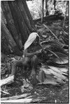 Pig festival, pig sacrifice, Tsembaga: woman, holding infant, flattens leaves for sacrifice house thatching