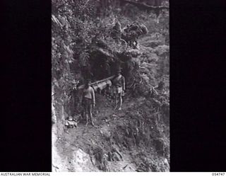 BULLDOG ROAD, NEW GUINEA. 1943-07-18. NATIVES CARRYING COMPRESSOR PARTS NEGOTIATE A NARROW AND DANGEROUS LEDGE ABOUT A MILE SOUTH OF ECCLESTON GAP. THIS LEDGE VARIES FROM EIGHTEEN INCHES TO TWO ..