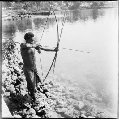 Man aiming a bow and arrow while standing on a riverbank, New Guinea, ca. 1936, 2 / Sarah Chinnery