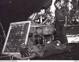 DUMPU, NEW GUINEA. 1943-10-15. SX27559 PRIVATE J. D. OLSON, MEDICAL ORDERLY OF THE 2/6TH AUSTRALIAN FIELD AMBULANCE NOTING PARTICULARS OF A PATIENT'S WOUNDS WHILE VX45728 DRIVER F. G. CLARKE ..