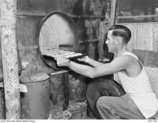 JACQUINOT BAY, NEW BRITAIN. 1945-04-26. PRIVATE W.J. MANALLACK, PLACING A TRAY OF TARTS INTO A HOME MADE OVEN MADE FROM A 44 GALLON DRUM AND CLAY. HE IS ATTENDING A SCHOOL OF COOKING CONDUCTED BY ..