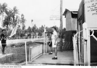 Two men stand at the main entrance to the RAN War Criminal Compound at the RAN shore base, as a bare footed member of the Royal Papuan Constabulary is about to raise the flag of the Royal Naval ..