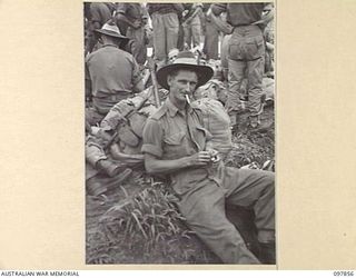 BORAM BEACH, NEW GUINEA. 1945-10-13. DRIVER D.J. COOMBS, LIGHTING A CIGARETTE WHILE WAITING FOR HIS TURN TO EMBARK FROM THE BEACHHEAD, IS ONE OF THE FIRST BATCH OF 6 DIVISION TROOPS TO LEAVE THE ..