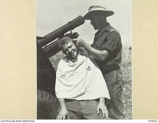 DALLMAN RIVER, NEW GUINEA, 1944-02-23. VX36006 GUNNER R. RYLEY (1), GIVES A HAIRCUT TO ANOTHER MEMBER OF THE 2/14TH FIELD REGIMENT, VX52963 GUNNER E.W. HILL (2), ALONGSIDE A 25-POUNDER GUN