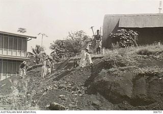 Japanese prisoners held at the RAN War Criminal Compound at an RAN shore base, using picks to remove a bank of dirt outside a building at the compound. The men are are being supervised by a member ..