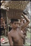 Mortuary ceremony: woman carries baskets partially filled with fiber skirts and banana leaf bundles