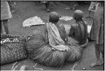 Mortuary ceremony, Omarakana: mourning women in long fiber skirts and necklaces, with heads shaved, seated beside banana leaf bundles