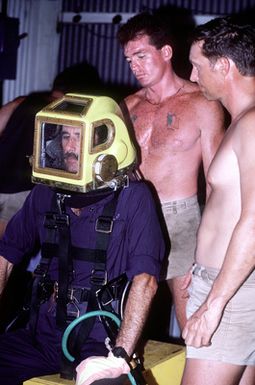 A Navy diver from Mobile Diving Salvage Unit One is fitted with a Mark 12 dive helmet in the dive platform workshop. The diver is preparing to study the wreck of the World War II Japanese merchant ship KIZUGAWA MARU as part of Project Sea Mark, a four-year undersea survey and mapping study of naval historic sites. The study is being conducted in conjuction with the U.S. Park Service's Submerged Cultural Resource Unit