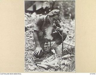 KARKAR ISLAND, NEW GUINEA. 1944-06-03. VX84603 SERGEANT B OSBORNE, 37/52ND INFANTRY BATTALION (1), LIGHTS HIS CIGARETTE FROM A FIRE BEFORE MOVING ON A PATROL