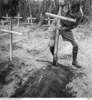 1942-12-16. GONA, PAPUA. CHURCH OF ENGLAND PADRE A.E. BEGBIE IN A SMALL BURIAL GROUND IN A CLEARING PUSHING A CROSS INTO THE GROUND AT THE HEAD OF ONE OF THE GRAVES OF SOLDIERS, MANY FROM 55/53RD ..