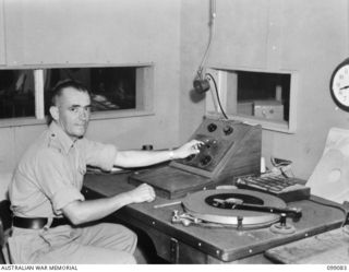 TOROKINA, BOUGAINVILLE. 1945-11-26. LIEUTENANT T. PEDRAZZINI, OFFICER COMMANDING, RADIO STATION 9AC TOROKINA, AUSTRALIAN ARMY AMENITIES SERVICE BROADCASTING STATION, AT WORK IN THE SMALL STUDIO