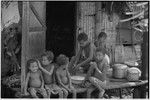 Children on a veranda, boy grates coconut into bowl, child (l) grooms younger child's hair, taro shoots (l) in background