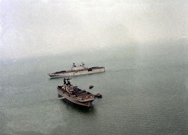 The passing of the amphibious assault ship USS NASSAU (LHA-4), in the foreground, and the amphibious assault ship USS SAIPAN (LHA-2) takes place during the turnover of Task Force 61 duties. NASSAU, the flagship for Marine Amphibious Ready Group (MARG) 1-82 relieves the SAIPAN, the flagship for MARG 3-81