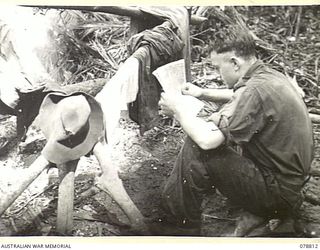 IDAKAIBUL, NEW GUINEA. 1945-01-25. QX37124 TROOPER K. CHERRY DRYING HIS WET CLOTHES OVER A CAMP FIRE WHILE READING THE LATEST ISSUE OF THE AUSTRALIAN ARMY NEWSPAPER, "GUINEA GOLD"