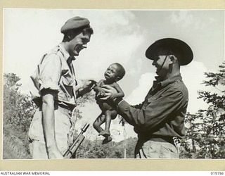 1943-06-29. NEW GUINEA. WAU- MUBO AREA. SGT. KEITH WITHERS, OF LEEDERVILLE, W.A., AND PTE. R. GUARD, OF CABAULTURIE, QUEENSLAND, WITH NAPON, BABY SON OF THE CHIEF OF ONE OF THE VILLAGES ON THE MUBO ..