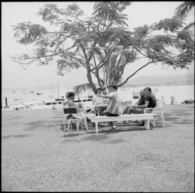 Six men socialising around a picnic area with sailing boats in the distance, Suva, Fiji, 1966 / Michael Terry