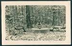 Man sits in forest on mill grinding stone, two machinery remnants sit on either side of grinding stone, relics from de Rays Expedition, New Ireland, New Guinea, 1930