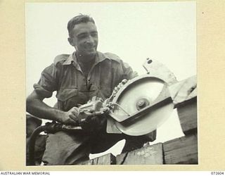 KILIGIA, NEW GUINEA. 1944-04-11. N190415 SAPPER J. TAYLOR, 8TH FIELD COMPANY, ROYAL AUSTRALIAN ENGINEERS USING A COMPRESSOR DRIVEN CIRCULAR SAW TO CUT LOGS FOR THE WOODEN DECKING OF A BRIDGE ACROSS ..