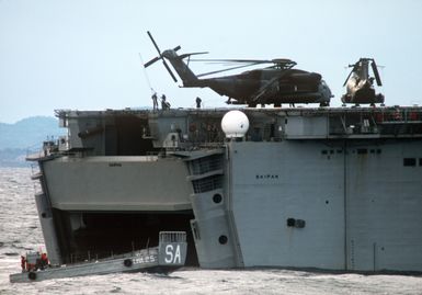 An LCM 6 mechanized landing craft exits from the well deck of the amphibious assault ship USS SAIPAN (LHA 2) during NATO Exercise NORTHERN WEDDING '86. On the flight deck are CH-46 Sea Knight and CH-53E Super Stallion helicopters