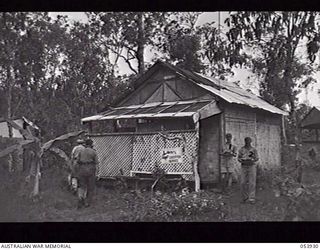 KOITAKI, NEW GUINEA. 1943-07-13. AUSTRALIAN ARMY EDUCATION SERVICE CENTRE AT SEVENTEEN MILE. VX118216 PRIVATE W. A. WARREN, IN CHARGE OF LIBRARY, STANDING (ON THE RIGHT) IN FRONT OF HUT WHILE ..