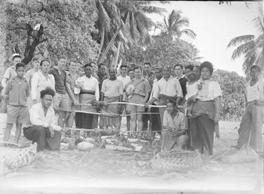 [Group of men and women gathered around spit roasting piglets outdoors]