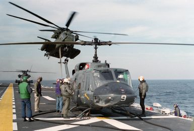 Flight deck crewmen perform a preflight inspection on a UH-1N Iroquois helicopter aboard the amphibious assault ship USS GUAM (LPH 9). A CH-53E Super Stallion helicopter is taking off in the background