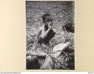 KIARIVU, NEW GUINEA, 1945-08-11. A NATIVE BOY SMOKING BY A FIRE. TROOPS OF 2/7 INFANTRY BATTALION ARE NOW IN THE AREA IN CONTROL OF THE EMERGENCY LANDING GROUND