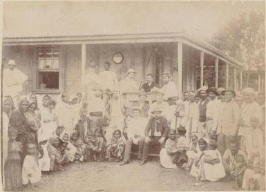 Indian labourers with their families and European overseers gathered in front of a house with verandah, Fiji, 1890