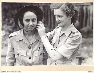 BRISBANE, QLD. 1945-02-21. PRIVATE B M WEBB, (1), HAS HER STEEL HELMET ADJUSTED BY PTE I V TESCH, (2). MEMBERS OF THE AUSTRALIAN ARMY MEDICAL WOMEN'S SERVICE, THEY AWAIT EMBARKATION FROM FRAZER'S ..