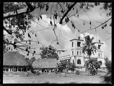 A three story stone [church?] with two local fale tele huts alongside, Apia, Western Samoa