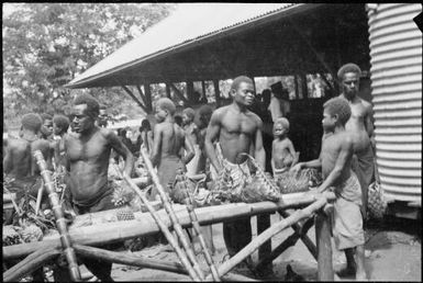 Men by a trestle table with a water tank to the right at the Boong, the native market, Rabaul, New Guinea, ca. 1929 / Sarah Chinnery