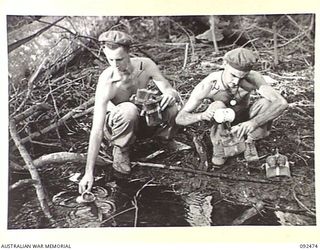 BOUGAINVILLE. 1945-05-22. CORPORAL R.W. HARTUNG (1) AND PRIVATE C.B. STEVENS (2), MEMBERS OF 24 INFANTRY BATTALION, FILLING WATER BOTTLES FROM A CREEK AT EGAN'S RIDGE ON THE BUIN ROAD SOUTH OF THE ..