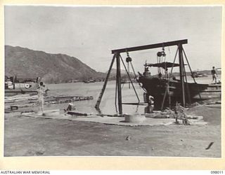 RABAUL, NEW BRITAIN. 1945-10-12. WHILE THE JAPANESE REPAIR ONE OF THEIR BARGES ON THE SLIPS AN AUSTRALIAN SOLDIER OF 55 PORT CRAFT COMPANY BATHES HIMSELF AT A WELL CLOSE BY