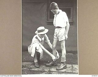 1943-03-15. NEW GUINEA. THIS PHOTO SHOWS SISTER VIDA DRYSDALE OF WEST WALLSEND N.S.W. SHOWING BILL HOW TO TIE THE LACES OF HIS BOOTS. BOOTS WERE SOMETHING UNKNOWN TO BILL. (NEGATIVE BY BOTTOMLEY & ..