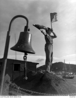 PORT MORESBY, PAPUA. C. 1944. A MEMBER OF THE RAAF RESCUE SERVICE SIGNALLING WITH SEMAPHORE FLAGS. IN THE FOREGROUND IS THE SHIP'S BELL SALVAGED FROM THE MV MACDHUI WHICH WAS THE TARGET OF ATTACKS ..