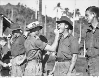 BOIKEN, NEW GUINEA. 1945-07-15. LIEUTENANT J.J. GARRICK, 2/6 CAVALRY (COMMANDO) REGIMENT (1), PLACING A TABLET IN THE MOUTH OF TROOPER G.E. MARTIN (2), DURING THE "ATEBRIN" PARADE